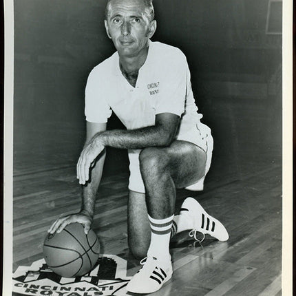 Black and white photo of basketball coach Bob Cousy sitting on a court with a ball, wearing a 'Cincinnati Royals' uniform.