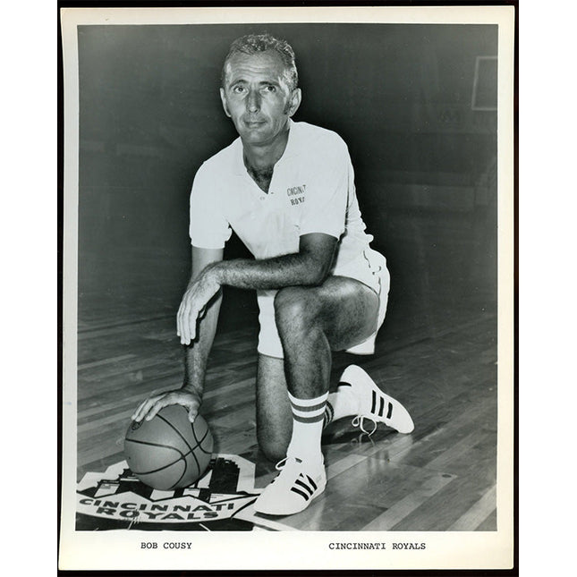 Black and white photo of basketball coach Bob Cousy sitting on a court with a ball, wearing a 'Cincinnati Royals' uniform.